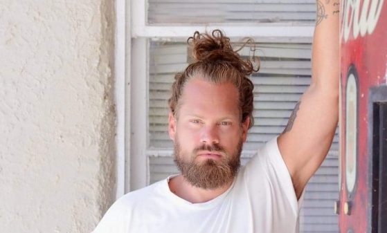 A photo of canadian musician Theo Tams leaning against a wall with a t-shirt on and his hair tied up